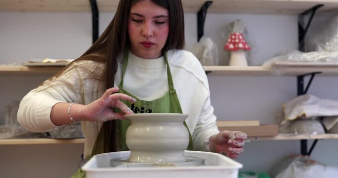 Concentrated young woman wearing a green apron carefully shaping a clay pot with her hands on a spinning pottery wheel, showcasing her artisanal skill in a creative workshop environment