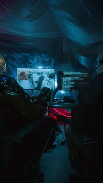 Vertical low key shot of cyberpunk programmers in smart visors fist bumping while monitoring surveillance footage and coding script on computers screens in dark office