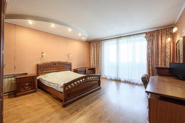 Fototapeta premium A cozy bedroom with a dark wooden bed, matching bedside tables, large window with sheer curtains, wooden desk, warm peach walls, and recessed ceiling lights.