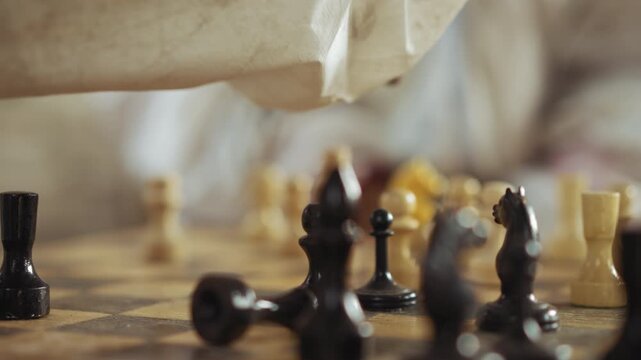 Side view close up of person in white sleeve reaching over wooden chessboard to pick up black piece during intense indoor match with scattered pieces and blurred background