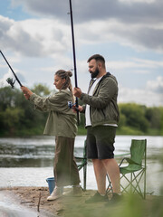 Young couple standing on the riverbank, holding fishing rods and enjoying a leisurely day outdoors,...