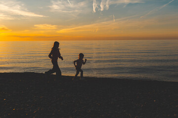 Two children running along pebble beach at sunset, silhouettes by calm sea, childhood freedom,...