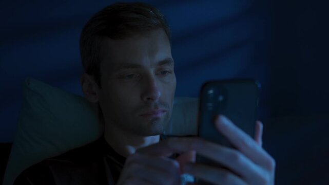 Sleepless young man browsing on his phone, doom scrolling in bed late at night lit by glowing blue screen light. Insomnia, procrastination or social media addiction. 