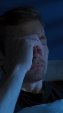 Young tired man with a phone in his hands scrolls through news feed or social media at night in his bedroom, but is interrupted by bad news or fatigue, disappointed and exhausted. Vertical video