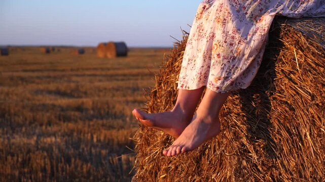 Barefoot woman in a long floral dress sitting on a round hay bale in a harvested field, gently swinging her legs in the warm evening light of sunset after a productive harvest day