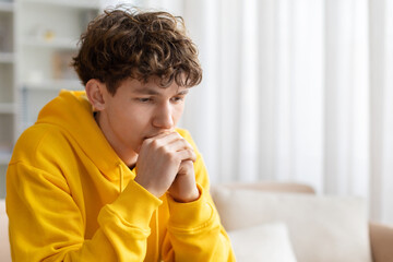 Thoughtful teenage boy sitting on sofa with hands near face, wearing yellow hoodie at home, deep in thought, worry, reflection, and quiet emotional moment indoors © Anastasiya