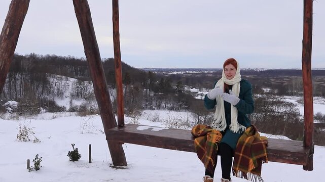 Beautiful woman sitting on a wooden swing in the middle of a winter hilly landscape. Peace, coziness, solitude and harmony with nature in winter.