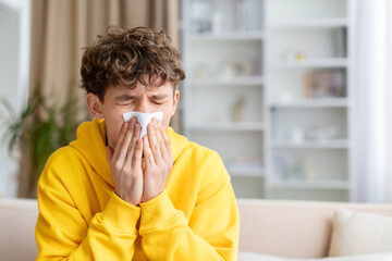 Sick teenage boy covering mouth with tissue while sneezing, sitting on couch at home, wearing yellow hoodie, cold and flu concept, health awareness indoors © Anastasiya