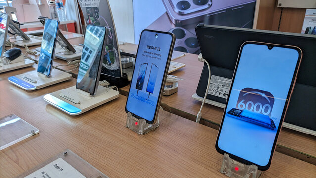 A smartphone display case in an electronics store, several Redmi phones are displayed on a wooden table with security stands and illuminated screens, showcasing the latest product features.