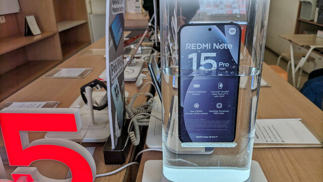 A smartphone display case in an electronics store, several Redmi phones are displayed on a wooden table with security stands and illuminated screens, showcasing the latest product features.