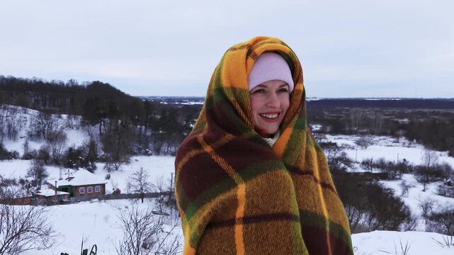 A young woman wrapped in a warm blanket stands against the backdrop of a snowy rural landscape. Winter peace, coziness, natural beauty and an atmosphere of solitude.