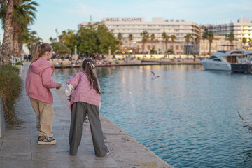Obraz premium Two eight year old girls feeding seagulls on waterfront at sunset with backlight, playful childhood outdoors