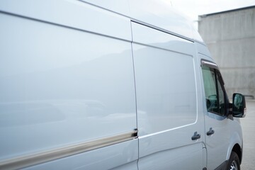 A clean white delivery van with a streamlined design is parked near a minimalist concrete building on a clear day with soft natural light reflecting off the vehicle's glossy surface
