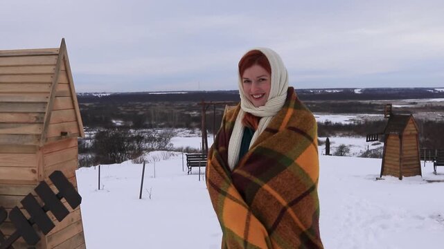A young woman wrapped in a warm blanket stands against the backdrop of a snowy rural landscape. Winter peace, coziness, natural beauty and an atmosphere of solitude.