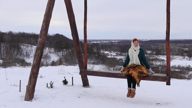 Beautiful woman sitting on a wooden swing in the middle of a winter hilly landscape. Peace, coziness, solitude and harmony with nature in winter.