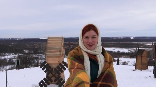 Beautiful girl in a warm coat and scarf runs with an open warm blanket in the middle of small wooden mills