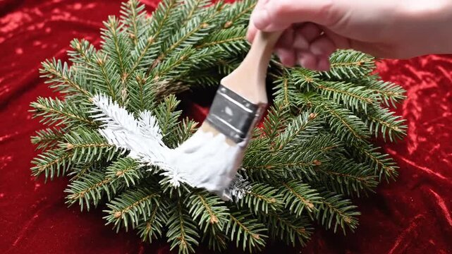 Cinematic slow motion capture of an artistic paintbrush decorating a festive evergreen pine wreath against a vibrant scarlet background, celebrating playful Christmas holiday craftsmanship.