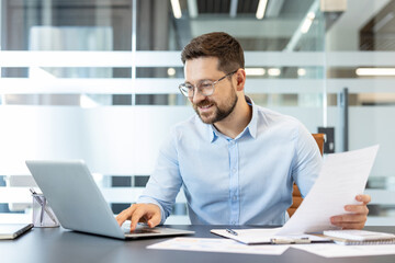 Young businessman engaged in professional work, holding paper document and typing on a laptop with...
