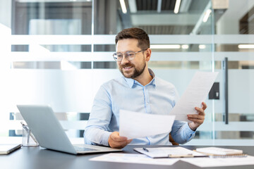 Fototapeta premium Professional bearded businessman wearing glasses analyzing documents and reviewing data while working on a laptop at a modern corporate office desk, demonstrating focus and business productivity