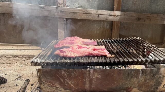 Authentic Argentine asado at a dusty construction site, featuring meat grilling on an improvised iron rack over a charcoal and wood fire with thick smoke