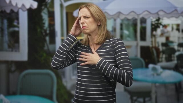 Woman touching ear then clutching chest in street cafe near outdoor tables and umbrellas, gingerly raising hands toward throat with pained expression; distress.