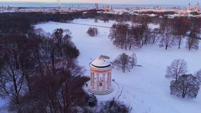 Historisches Zentrum von Muenchen im Winterschnee. Drohnenpanorama vom Englischen Garten zum alten Stadtkern mit schneebedeckten Daechern und dem Monopteros im goldenen Licht