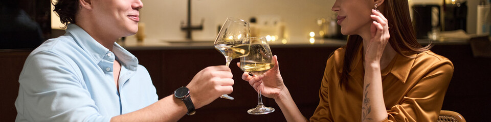 Header of young Caucasian man and young Caucasian woman sitting together clinking wine glasses, smiling and engaging in conversation, enjoying evening indoors at restaurant or bar