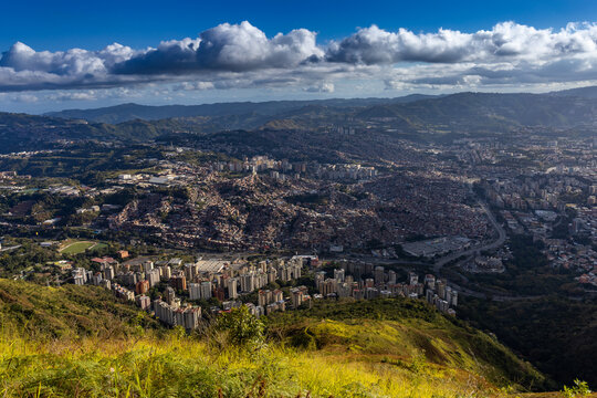 Wide panoramic view of the Caracas valley from PGP Galindo in Waraira Repano National Park, showing the dense urban sprawl of Petare and the eastern residential districts under a cloudy blue sky
