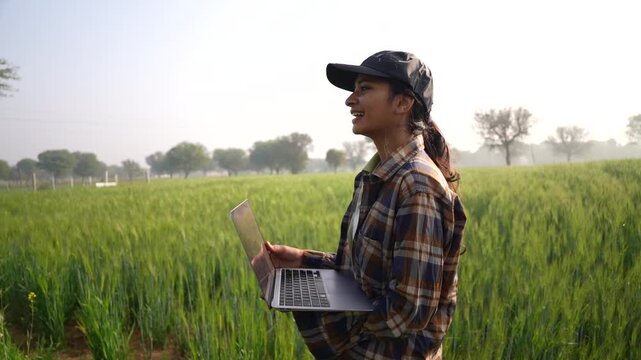 Young woman using laptop on agricultural field. Business lady pointing her finger and looking at notebook screen. Modern female farmer wearing cap and inspecting the wheat green field crop.
