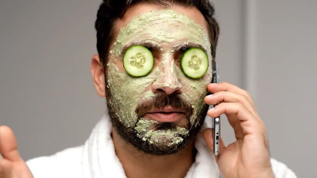 A man in a bathrobe with a face mask and cucumber slices on his eyes is talking on a phone