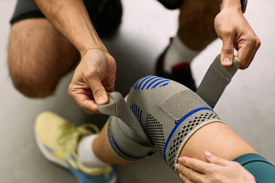 Caucasian young adult man assisting Caucasian young adult woman by wrapping elastic bandage around knee during physical therapy rehabilitation session focusing on injury recovery