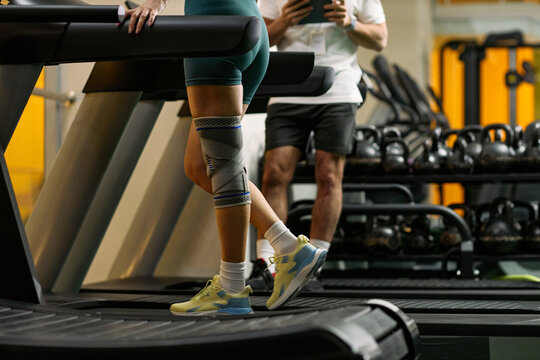 Caucasian young adult woman walking on treadmill with knee brace during physical therapy session in rehabilitation center, male therapist standing nearby holding digital tablet