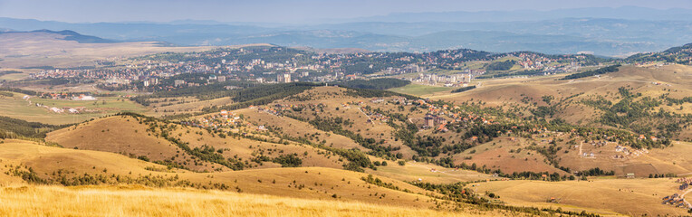 Obraz premium Panoramic view of Zlatibor landscape, featuring golden rolling hills, sparse vegetation, and a distant town under a pale sky