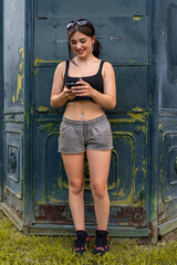 Young woman standing by a distressed wall, smiling while typing on her smartphone