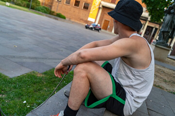 Young man sitting on ground in bucket hat and tank top observing the street and police car