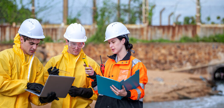 Team of environmental scientists in hazmat suits using laptop and discussing water monitoring data near industrial fuel storage tanks. Three professionals inspecting water contamination at site