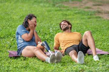 Fototapeta premium True friendship and equality happy woman with prosthetic leg relaxing on yoga mat with her supportive friend in park. Smiling female amputee with artificial limb enjoying with companion on grass.