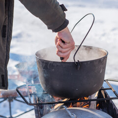 Person preparing hot food outdoors during winter using a metal cauldron over open fire. Close view of hands cooking a meal in cold weather