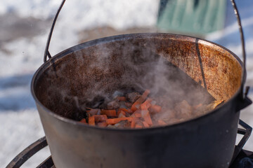 Person preparing hot food outdoors during winter using a metal cauldron over open fire. Close view of hands cooking a meal in cold weather