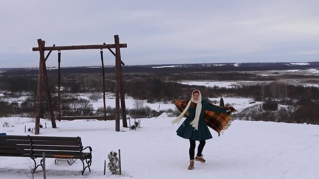 A beautiful girl in a warm coat and scarf runs with an open warm blanket on a snowy mountain with a beautiful panorama of valleys and forests, enjoying freedom, winter and nature.