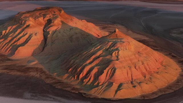 Aerial view of a rugged mountain terrain with layered rock formations, bathed in warm sunlight against a backdrop of desert, Karynzharyk, Kazakhstan.