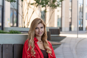 Fototapeta premium Portrait of Blonde Woman in a Red Shirt Sitting on a Bench in City Street