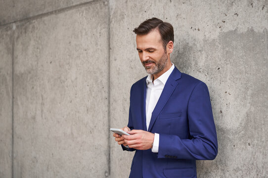 Adult businessman using mobile phone leaning against concrete wall