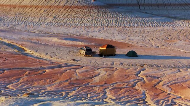 Aerial view of a vehicle and tents pitched on the wavy, multi-toned landscape, creating a striking contrast of colors and textures, Karynzharyk, Mangystau Region, Kazakhstan.