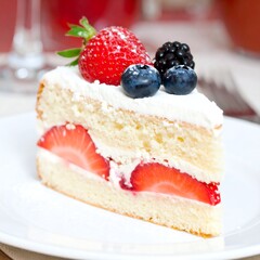 A slice of frosted cake with fresh strawberries and berries, presented on a white plate, against a blurred backdrop