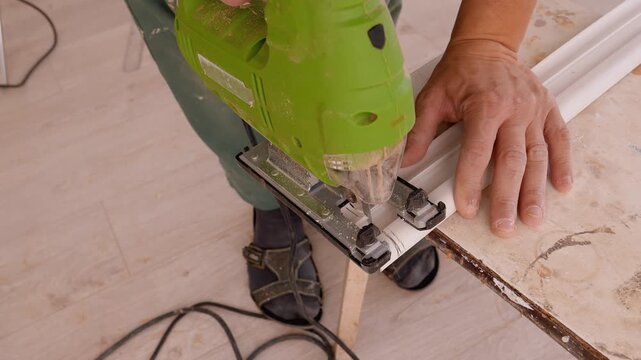 Skilled worker who works with an electric puzzle to accurately cut a piece of white plastic ceiling skirting board during home renovation looking from a high angle