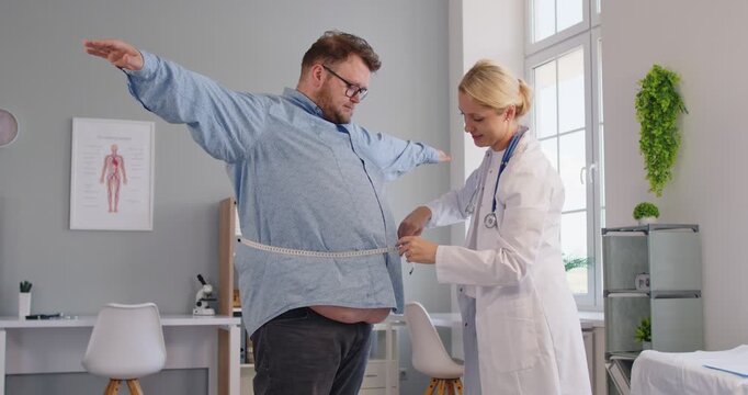 Clinician helps an overweight fat patient in a clinic exam, using tape to record waist measurement and provide obesity counseling on diet and health risks. Nutritionist measures patient waist