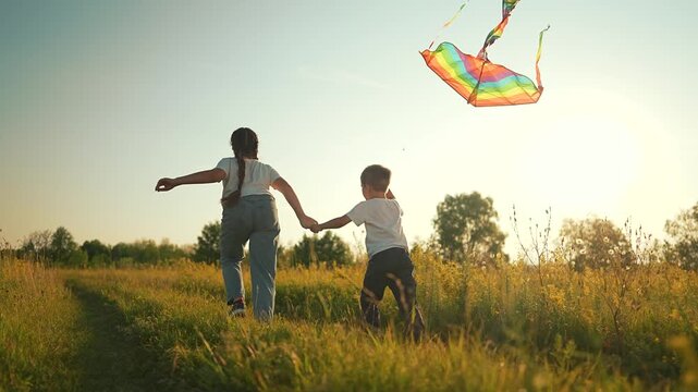 Girl and boy run holding hands watching rainbow kite in meadow at sunset outdoors. Sister with brother plays using colorful kite. Children run in field. Girl and boy watch kite. Siblings in meadow.