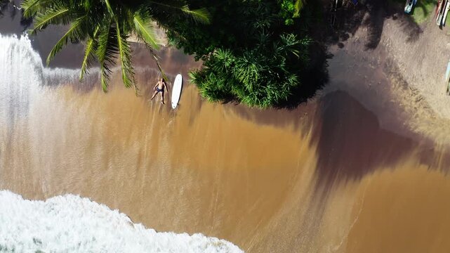 4K top view aerial shot of lonely tired surfer man lying in star pose on sandy beach washed by ocean waves with surfboard. Happy summer vacation and active sporty lifestyle concept. Mirissa, Sri Lanka