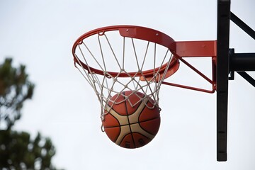 Naklejka premium Classic orange basketball falling through net of red rim against clear sky background view